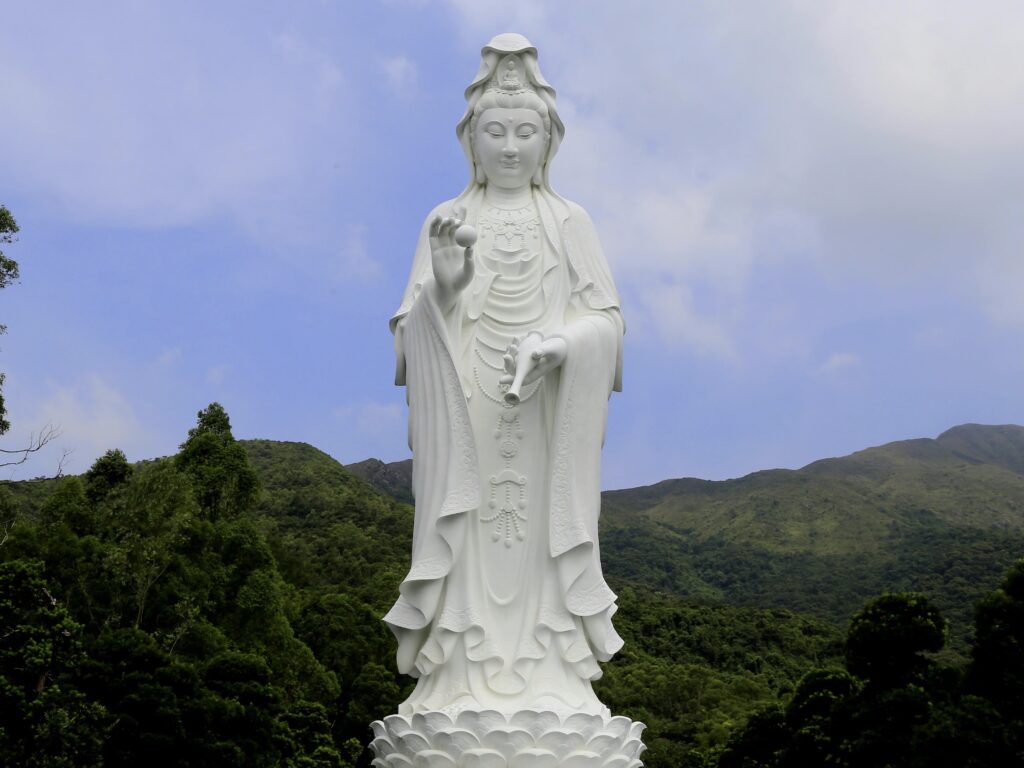 White statue of Kuan Yin, the Buddhist goddess of compassion, standing in nature against a blue sky.