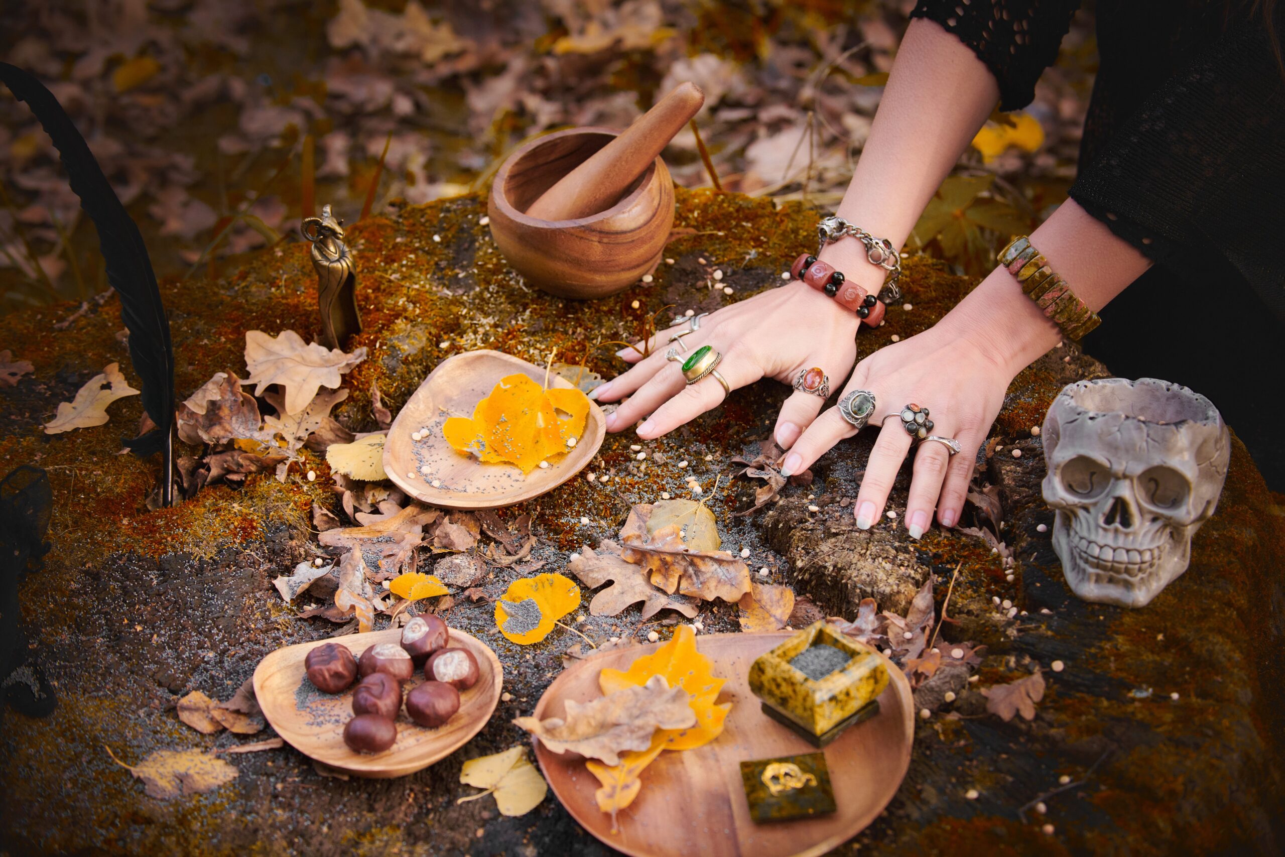 Autumn Ritual - Plant Wisdom and The Descent Woman’s hands adorned with jewelry resting on a mossy rock surrounded by autumn leaves, a skull, a small witch figurine, nuts, and a wooden mortar and pestle arranged for a seasonal ritual.